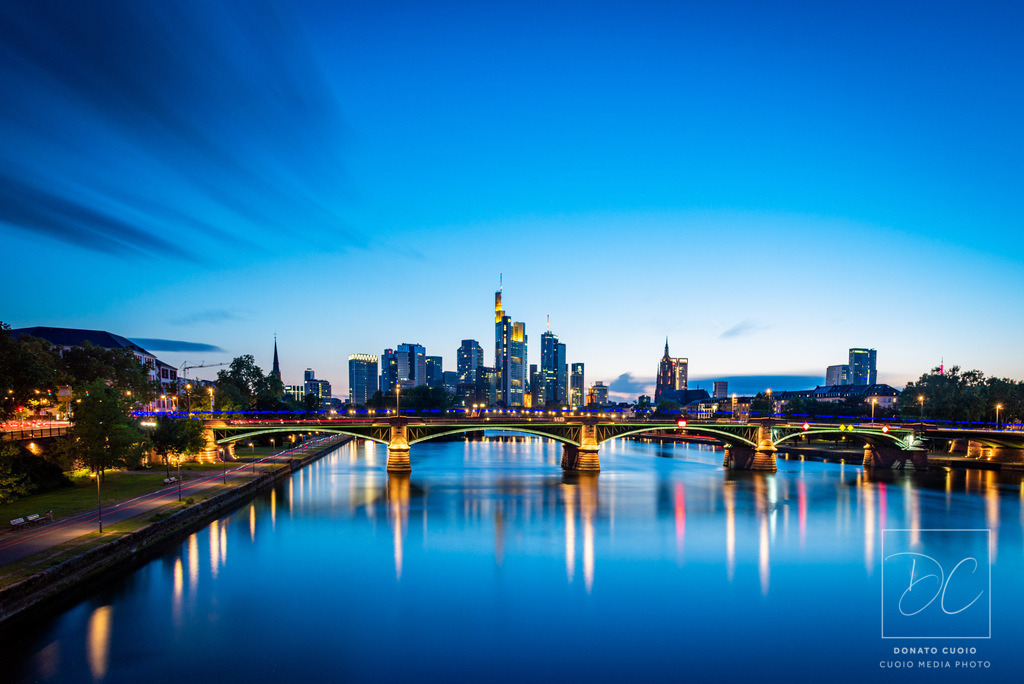Frankfurt – Blue Hour Serenity | Die Stadt in ihrer elegantesten Stunde: Die Blaue Stunde schenkt Frankfurt eine fast magische Tiefe. Spiegelungen im Wasser, goldene Lichter auf der Brücke, sanfte Bewegungen der Wolken – ein beruhigendes und zugleich kraftvolles Wandmotiv.Frankfurt Skyline blaue Stunde mit Langzeitbelichtung und Lichtreflektionen im Main - Realisiert mit Pictrs.com