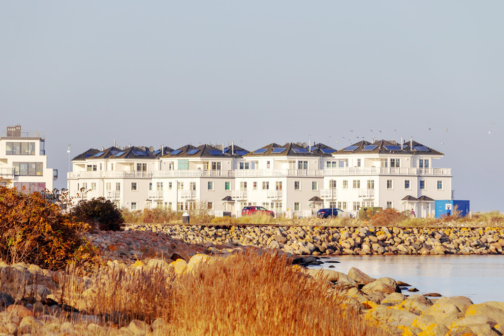 Wandbild: Ferienwohnungen am Meer in Olpenitz | Dieses Wandbild im Querformat zeigt Ferienwohnungen am Meer in Olpenitz im Herbst. Die abendliche Sonne taucht alles in einen leichten Rotton. Im Vordergrund befindet sich Strandhafer sowie eine Steinmole.  - Realisiert mit Pictrs.com