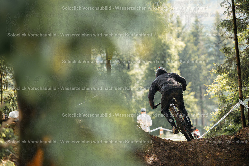 Mountainbike-Action im Wald | Ein Mountainbiker in voller Ausrüstung, einschließlich Helm und Schutzbrille, springt über eine Rampe in einem bewaldeten Gelände. Der Hintergrund besteht aus dichtem Grün und Bäumen, was eine natürliche und abenteuerliche Atmosphäre schafft. Ein weiterer Mann steht am Rande der Rampe und beobachtet den Mountainbiker. - Realisiert mit Pictrs.com