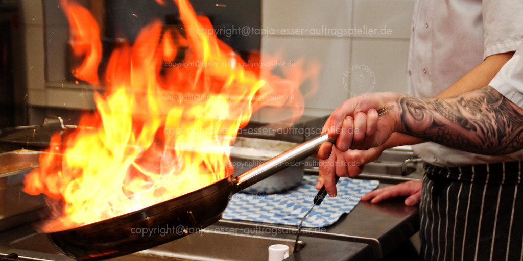 gastronomie_d | Authentische Nahaufnahme eines männlichen Kochs, der eine Pfanne schwenkt in der er Fleisch flambiert. Auf dem Bild sind zwei Köche in einer gastronomischen Küche am Herd zu sehen. 