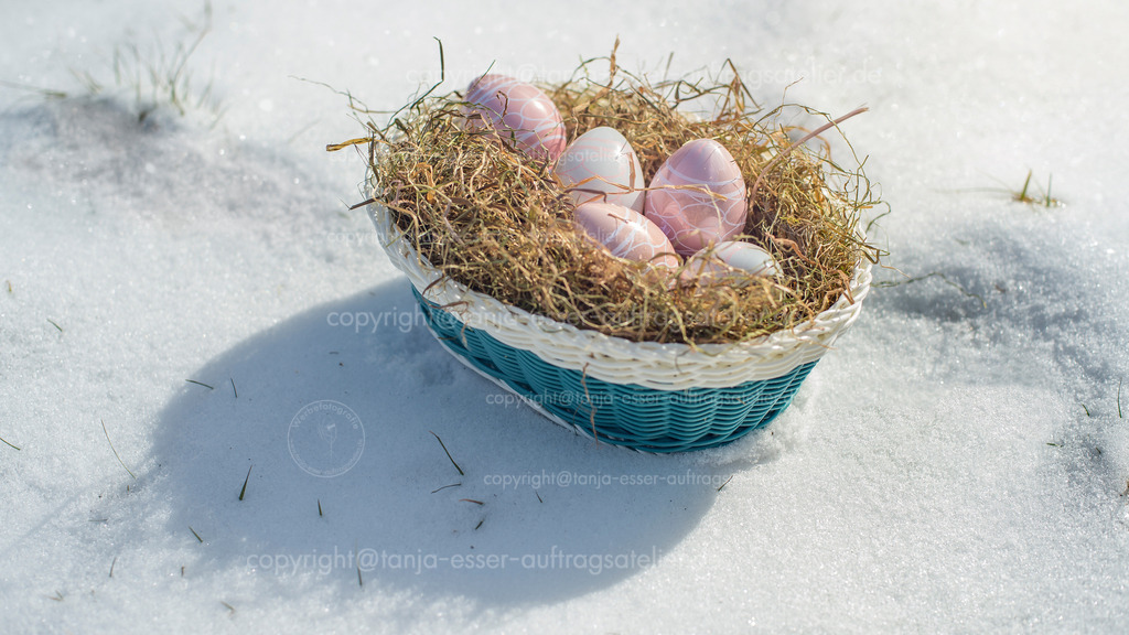 Osternest steht auf verschneiter Wiese. Aufnahme von oben. | Eine Wiese ist mit Schnee bedeckt. Ein natürliches Nest aus Heu enthält weiß und rosa schimmernde Ostereier aus Glas. Copy Space. Hintergrund für verschneite Osterfeiertage. 