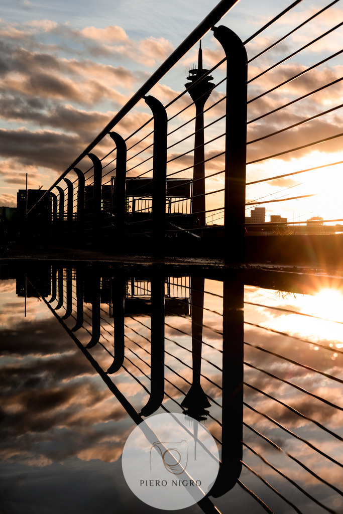 Rheinufer und Rheinturm Düsseldorf: Eine atemberaubende Reflektion bei Sonnuntergang | DE: Diese Fotografie zeigt das Rheinufer Düsseldorf nach einem Regenschauer bei Sonnenuntergang. Die Silhouette des Rheinturms reiht sich ein in die Silhouette des Geländers und erzeugt eine fast perfekte Spiegelung mit dem Regenwasser auf dem Boden.

EN: This photograph shows the banks of the Rhine in Düsseldorf after a rain shower at sunset. The silhouette of the Rhine Tower joins the silhouette of the railing and creates an almost perfect reflection with the rainwater on the ground.
 - Realisiert mit Pictrs.com
