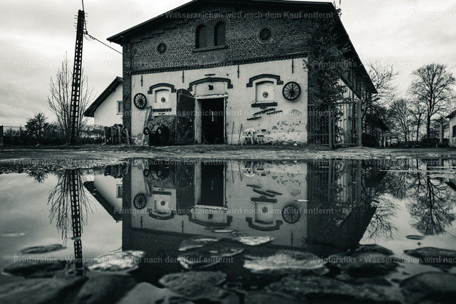 Altes Gebäude mit Spiegelung | Schwarzweißaufnahme eines alten Gebäudes, das sich in einer großen Wasserfläche spiegelt. Die ruhige Symmetrie, die verwitterte Fassade und die reduzierte Farbwirkung erzeugen eine stille, grafische und leicht melancholische Atmosphäre. - Realisiert mit Pictrs.com