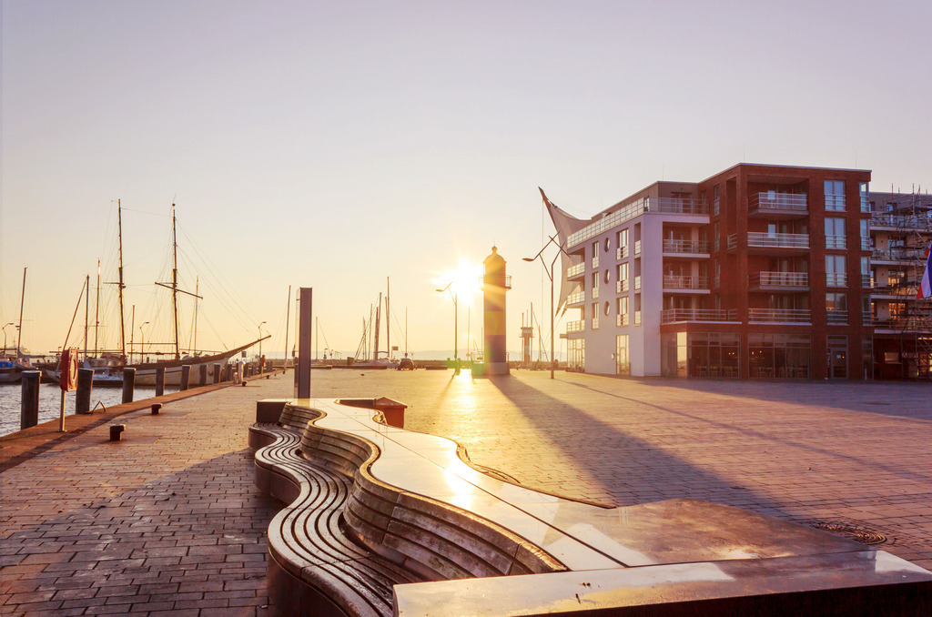 Wandbild: Sonnenschein am Ostseehafen | Dieses Wandbild zeigt eine schöne Morgenstimmung am Ostseehafen. Die tiefstehende Sonne steht direkt hinter einem kleinen Leuchtturm.Durch die morgendlichen Lichtstimmung ergeben sich lange Schatten. Im Hintergrund auf der linken Seite befindet sich ein Traditionssegler.  - Realisiert mit Pictrs.com
