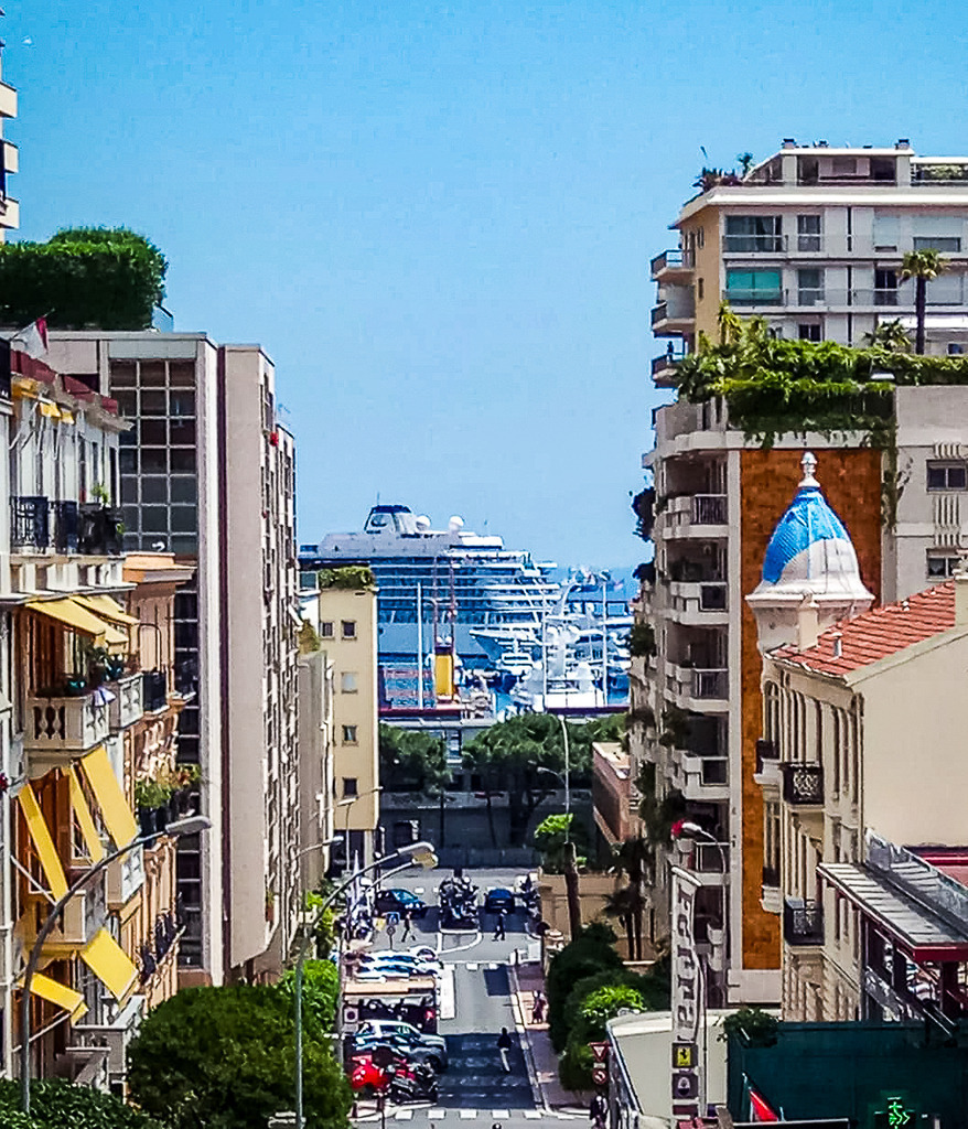 Wandbild Kunstdruck Poster – Monaco Street View: Luxus & Meer an der Côte d'Azur von Robert Steinke | Hol dir das unvergleichliche Lebensgefühl der französischen Riviera direkt in deine vier Wände. Diese beeindruckende Street-View-Fotografie aus Monaco fängt die exklusive Atmosphäre des Fürstentums in Perfektion ein. Der Blick wandert über die charakteristischen, mediterranen Häuserfassaden und sattgrünen Palmen hinunter zum tiefblauen Himmels-Ozean, auf dem ein majestätisches Kreuzfahrtschiff im Hafen verweilt. Das warme, goldene Sonnenlicht und die maritime Weite versprühen pure Eleganz und Urlaubsflair.Dieses luxuriöse Motiv ist der perfekte architektonische Blickfang für ein modernes Wohnzimmer, das stilvolle Büro, ein exklusives Loft oder für eine sommerliche, inspirierende Stimmung im Schlafzimmer. Es bringt Licht, Weite und den Glanz der Côte d’Azur in jeden Raum.Um die strahlenden Farben des Meeres und die feinen Details der Architektur perfekt zur Geltung zu bringen, empfehle ich den Druck auf unseren High-End-Materialien: Erlebe kinoreife Tiefenwirkung auf edlem Acrylglas, modernste Eleganz auf robustem Alu-Dibond oder klassischen Galerie-Charakter auf einer matten Leinwand. Ein zeitloses Meisterwerk für Reise- und Kunstliebhaber! - Realisiert mit Pictrs.com