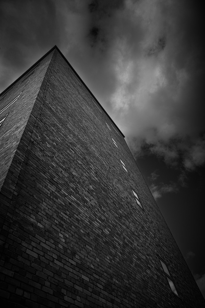 Dark Urban Geometry – Minimalist Architecture Perspective | This fine-art architectural photograph captures the dramatic perspective of a modern brick building rising into a dark and textured sky.The strong geometric lines and the upward view create a powerful visual composition that emphasizes structure, minimalism, and urban atmosphere. The contrast between the dark bricks and the moving clouds adds tension and depth to the scene.This image is ideal as modern wall art for living spaces, offices, architecture studios, and minimalist interiors. - Realisiert mit Pictrs.com