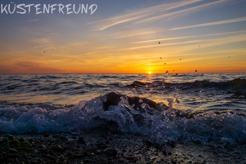 Sonnenuntergang am Meer // Fotoserie Sunset & Splash | Ein Sonnenuntergang am Meer, eingefangen aus meiner Fotoserie „Sunset & Splash“. Sanfte Wellen umspülen einen Findling und erzeugen zarte Gischt. Im Vordergrund zeichnet sich der Spülsaum ab, der an diesem Naturstrand überwiegend aus Steinen besteht. Am Horizont versinkt die Sonne langsam und taucht die Szene in das warme Licht der goldenen Stunde, die sich sanft dem Ende zuneigt.<br><br><strong>Tipp:</strong> Statt das klassische Querformat zu wählen, kannst du dieses Sonnenuntergangsbild am Meer auch ganz flexibel zuschneiden – ob als quadratisches Wandbild, Hochkantformat oder Panorama. So passt dein persönliches Ostsee Bild perfekt in jeden Raum und wird zum individuellen Highlight an deiner Wand.