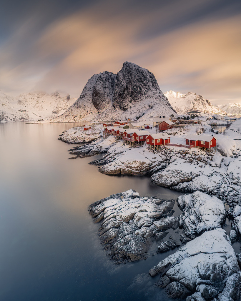 Norwegisches Fischerdorf vor verschneitem Berg | Ein kleines Fischerdorf liegt auf felsigen Inseln vor einem steil aufragenden, verschneiten Berg. Das ruhige Wasser und die sanften Wolkenbewegungen verleihen der Landschaft eine stille, nordische Wirkung. - Realisiert mit Pictrs.com