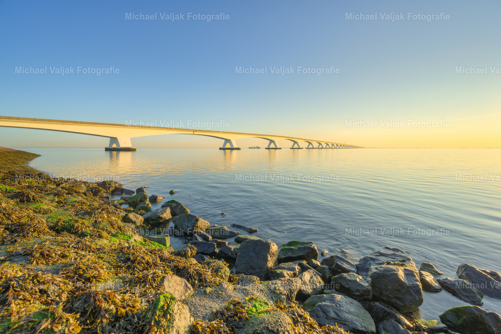 Zeelandbrücke im goldenen Morgenlicht | Die ersten Sonnenstrahlen des Tages lassen die imposante Zeelandbrücke in warmem, goldenem Licht erstrahlen. Vom Ufer aus fällt der Blick über die markanten Steine im Vordergrund hinüber zur langen Reihe der Brückenpfeiler, die sich weit über das Wasser erstreckt.Das natürliche Morgenlicht bringt die Strukturen der Steine, des Wassers und der Brücke eindrucksvoll zur Geltung. Die ruhige Küstenlandschaft und der warme Glanz der aufgehenden Sonne schaffen eine klare und zugleich stimmungsvolle Atmosphäre.Ein authentisches Landschaftsfoto aus Zeeland, Niederlande, das die besondere Schönheit eines frühen Morgens an der niederländischen Küste einfängt. - Realisiert mit Pictrs.com
