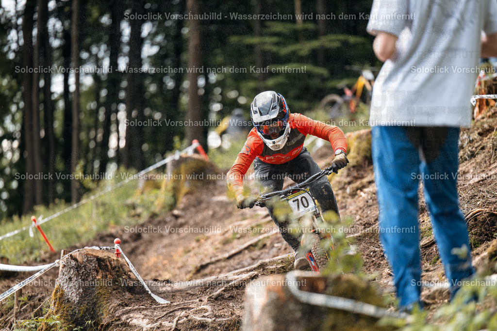 Mountainbike-Action | Ein Mountainbiker in voller Ausrüstung rast mit hoher Geschwindigkeit einen staubigen, kurvigen Trail hinunter. Er trägt einen roten Helm, eine dunkle Brille und eine schwarze Jacke mit roten Akzenten. Er trägt auch Handschuhe und eine Nummer 70 auf dem Rücken. Der Trail ist mit Wurzeln und Steinen übersät, und es gibt einige Baumstümpfe am Rand. Ein Zuschauer steht am Rand des Trails und beobachtet den Mountainbiker. - Realisiert mit Pictrs.com