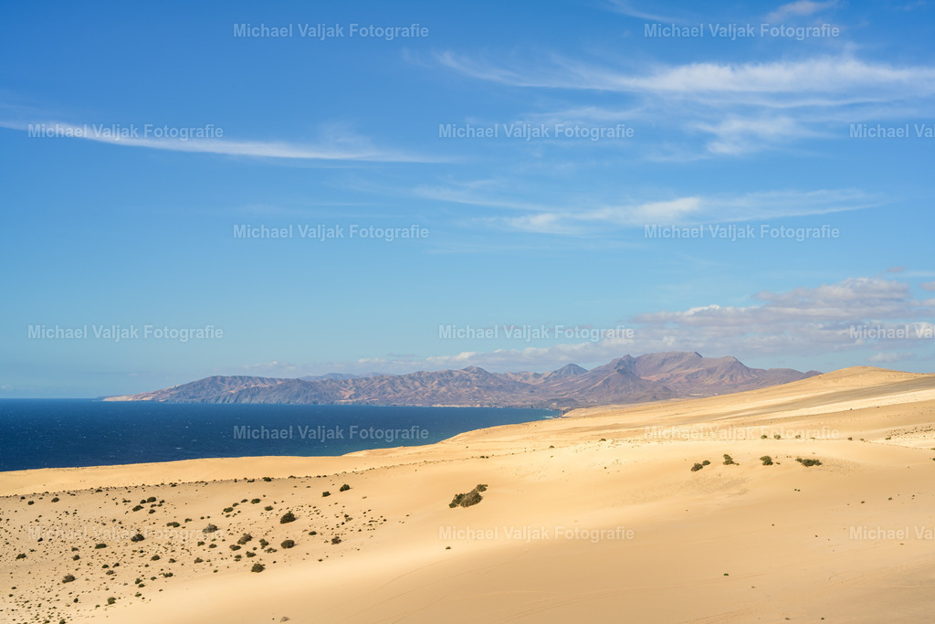 Blick über die Dünen bei Jandía auf Fuerteventura | Der Blick über die Dünen des Istmo de La Pared zeigt ein ruhiges Spiel aus Licht und Schatten, das sich über die weichen Sandflächen zieht. Die Sonne hebt einzelne Linien und Kanten hervor, während andere Partien in ein kühleres Halbdunkel fallen. In der Ferne steigen die Formen des Montaña Cardón klar und markant aus der Landschaft auf. Ein Moment, in dem Struktur, Tiefe und die stille Weite des Südens ineinander übergehen. - Realisiert mit Pictrs.com