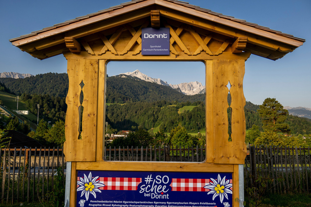 Bilderrahmen mit Alpenaussicht: So schee bei Dorint | Ein aufwendig gezimmerter Holzrahmen des Dorint Sporthotels in Garmisch-Partenkirchen setzt die beeindruckende Kulisse der bayerischen Alpen gekonnt in Szene. Der Blick wandert durch das Holzfenster direkt auf sattgrüne Bergwälder und die dahinter aufragenden, sonnenbeschienenen Massive: Die markante Pyramide der Alpspitze, der wilde Jubiläumsgrat und die imposante Zugspitze präsentieren sich wie in einem lebendigen Gemälde. Liebevolle Details wie das Edelweiß-Dekor und der Spruch „#soscheebeidorint“ wecken echte Urlaubsfreude und laden zum Innehalten ein. - Realisiert mit Pictrs.com