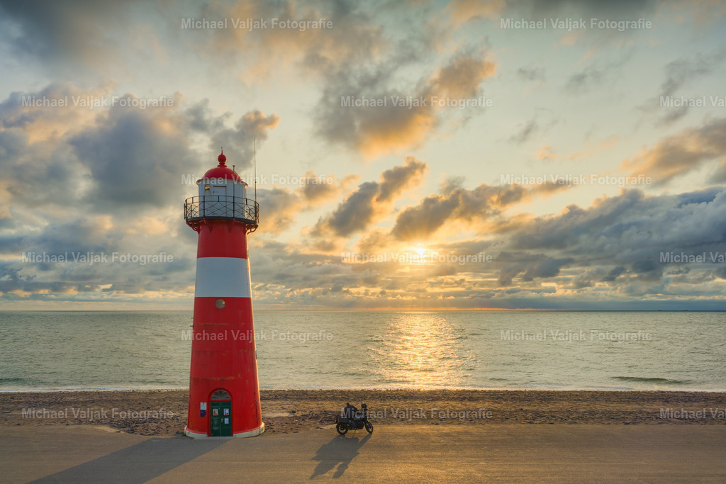 Rast am Kleinen Leuchtturm Westkapelle | Kurz vor Sonnenuntergang zeigt sich der Kleine Leuchtturm von Westkapelle in warmem, stimmungsvollem Licht. Der markante Turm erhebt sich vor dem weiten Himmel und der ruhigen Küstenlandschaft von Zeeland.Am Fuß des Leuchtturms hat ein Motorradfahrer eine kurze Rast eingelegt. Das Motorrad und die kleine menschliche Szene verleihen dem ansonsten weitläufigen Küstenmotiv eine besondere Lebendigkeit und erzählen ihre ganz eigene Geschichte.Ein atmosphärisches Landschaftsfoto von Westkapelle in Zeeland, Niederlande, das die besondere Verbindung von Meer, Küste, Architektur und Reisefreiheit einfängt. - Realisiert mit Pictrs.com
