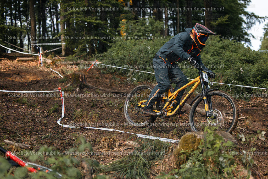 Mountainbike-Action | Ein Mountainbiker in voller Ausrüstung rast einen steilen, erdigen Hang hinunter. Er trägt einen schwarzen Helm mit orangefarbenen Akzenten, eine dunkle Brille und eine schwarze Jacke mit orangefarbenen Details. Sein Mountainbike ist mit goldenen Akzenten versehen. Der Hintergrund besteht aus dichtem Laubwerk und einem Netzwerk aus weißen Markierungen, die wahrscheinlich Teil eines Mountainbike-Rennstrecken sind. - Realisiert mit Pictrs.com