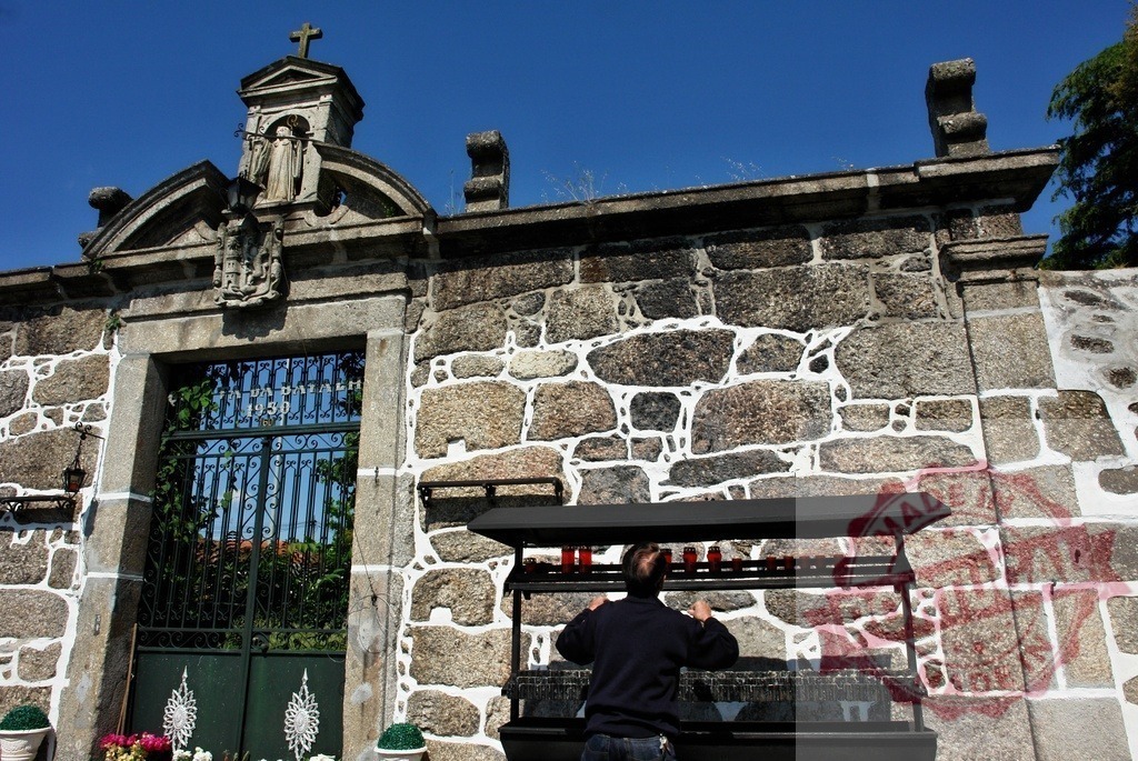 Man lighting votive candles at religious shrine entrance. Region Norte - Portugal | Man placing prayer candles at a stone wall entrance with architectural details and clear sky - Realized with Pictrs.com