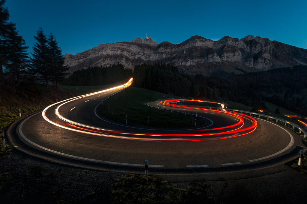 Kurvenlicht – Nachtfahrt am Säntis | Die geschwungene Straße am Fuße des Säntis wird in der Dämmerung von roten und weißen Lichtspuren durchzogen. Durch die Langzeitbelichtung treffen alpine Landschaft auf dynamische Bewegung – ein Bild voller Kontraste und Energie. - Realisiert mit Pictrs.com
