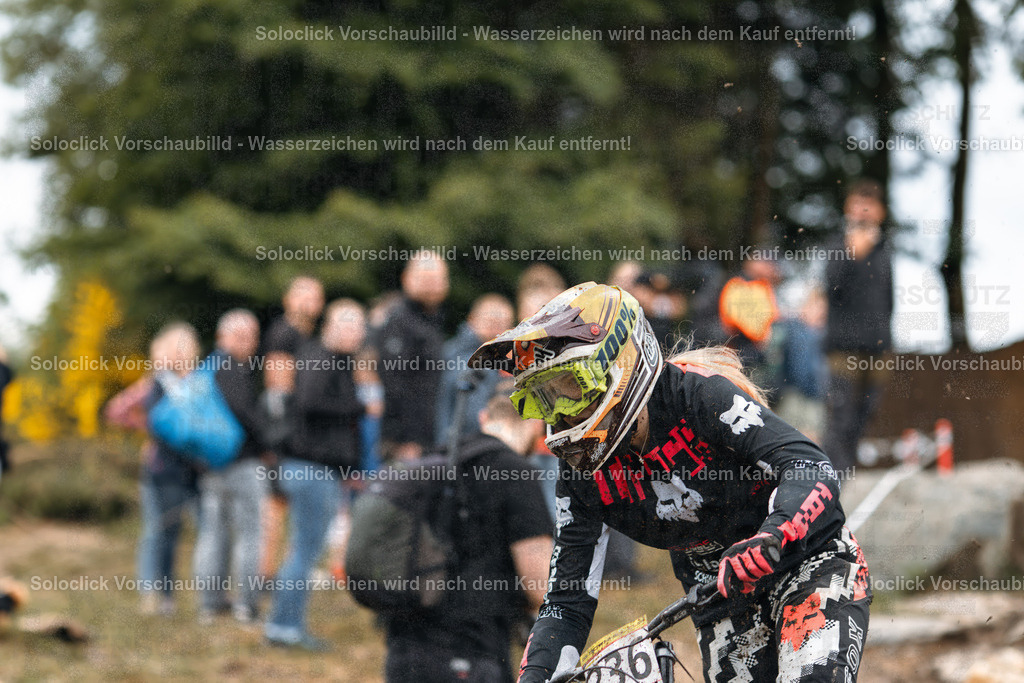 Mountainbike-Action im Gelände | Das Foto zeigt einen Mountainbiker, der in voller Ausrüstung durch einen schlammigen Bereich fährt. Er trägt einen Helm, eine Brille und spezielle Kleidung, die ihn vor Schmutz und Verletzungen schützen sollen. Im Hintergrund sind mehrere Zuschauer zu sehen, die das Rennen beobachten. Die Szene wirkt dynamisch und actionreich. - Realisiert mit Pictrs.com