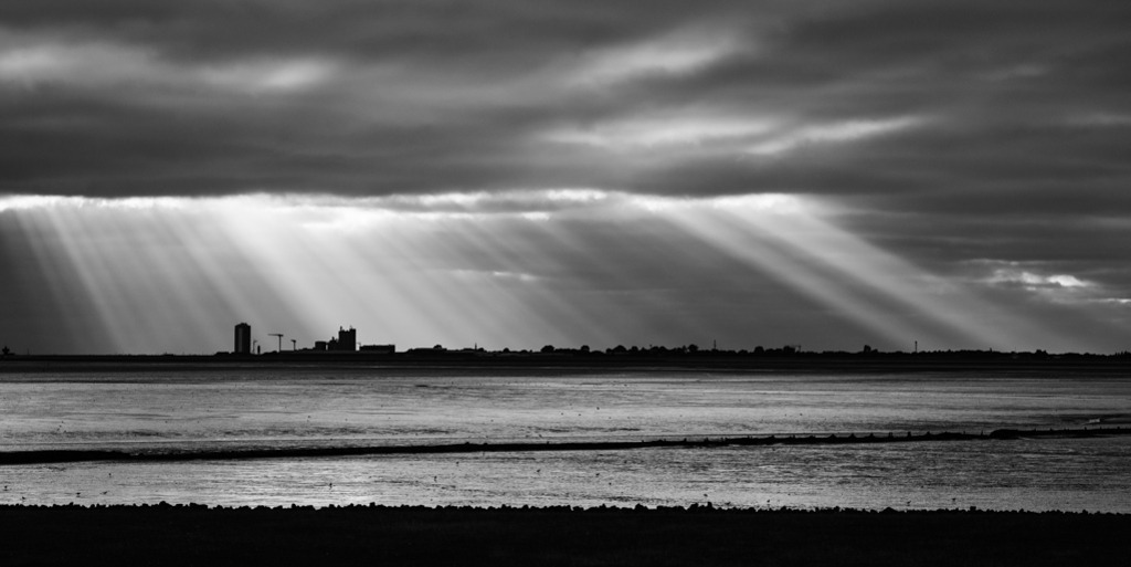 Spektakel über Büsum | Das Wattenmeer der Nordsee bei Ebbe. Durch eine schwere Wolkendecke bricht vereinzelt das Licht, entfacht ein Spektakel aus Lichtstrahlen über der Silhouette von Büsum. — Auflösung des Originals: 8256 x 4128 px. - Realisiert mit Pictrs.com