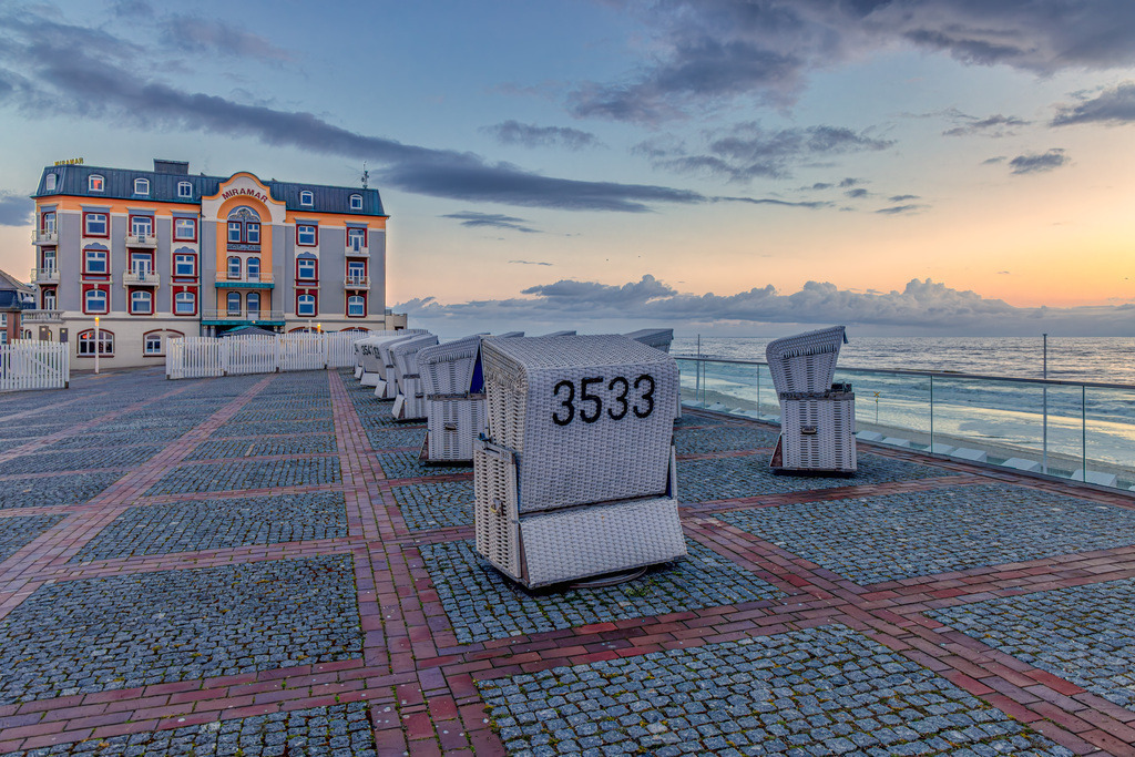 Abendliche Stimmung am Grand Hotel Miramar in Westerland auf Sylt | Hotel Miramar: Das markante Gebäude im Jugendstil auf der linken Seite ist das renommierte Hotel Miramar, ein geschichtsträchtiges 5-Sterne-Luxushotel direkt an der Westerländer Promenade.Typische Strandkörbe: Im Vordergrund stehen mehrere für die deutsche Nordseeküste charakteristische Strandkörbe auf der gepflasterten Promenade, einer davon trägt die gut lesbare Nummer 3533.Strandpromenade: Der gepflasterte Bereich ist Teil der Kurpromenade, die einen direkten Blick auf das offene Meer und den darunter liegenden Sandstrand bietet. - Realisiert mit Pictrs.com