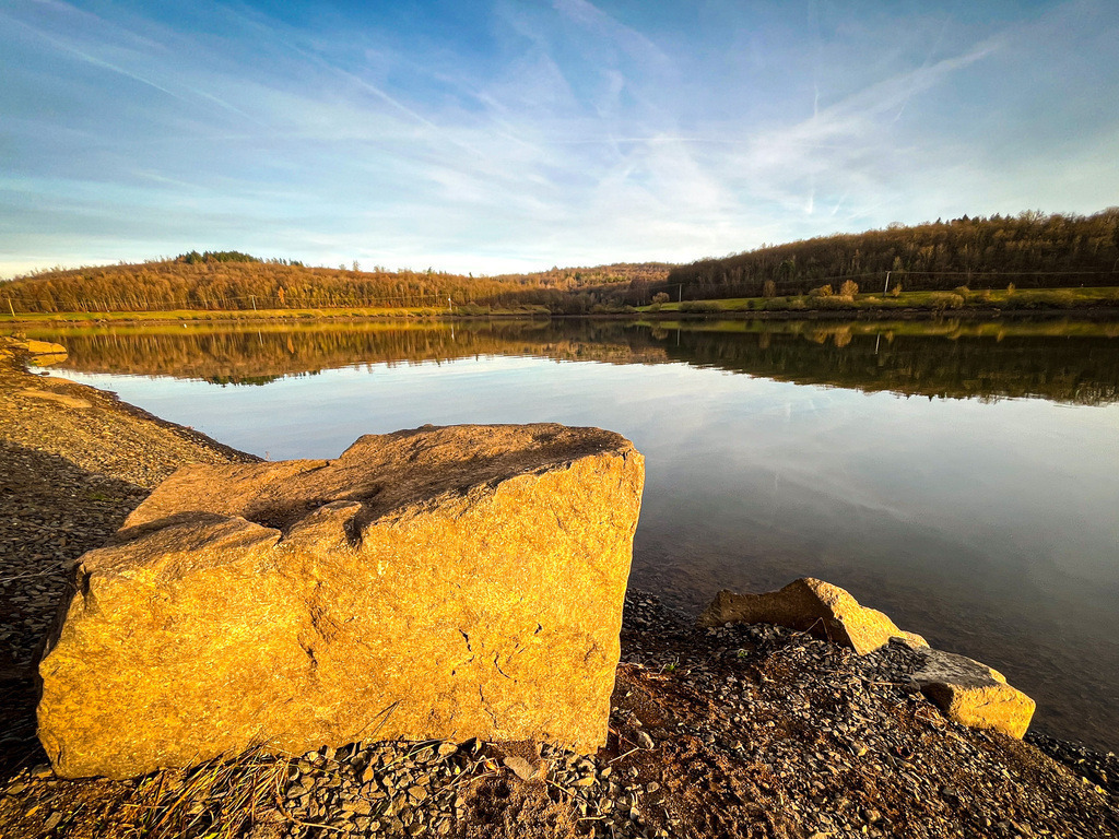 Steinsee - Schotten | Direkt am Uferrand lädt ein massiver Stein im Licht der Abendsonne zum Verweilen ein.Der Nidda-Stausee schafft im Dezember eine Atmosphäre der Gegensätze. Während die Bäume bereits ihr Blätterkleid für den Winter abgelegt haben, wirkt das Gras grün und üppig. Während am Horizont die heimischen Wälder zu sehen sind, scheint das unmittelbare Seeufer fremd, steinig und kahl. Zusammen schaffen diese Eindrücke eine einzigartige Kulisse, die auch abseits der Badesaison einen Besuch wert ist. - Realisiert mit Pictrs.com