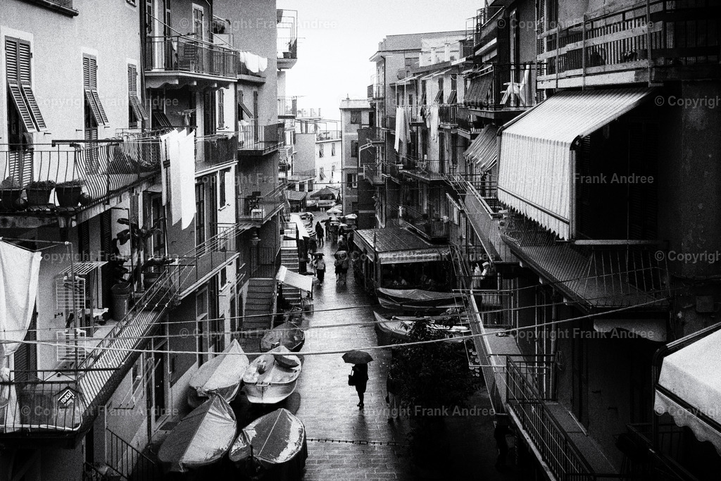 Regen in Manarola | In der Altstadt von Manarola Cinque Terre steht eine Frau mit Regenschirm neben abgestellten Fischerbooten. Street Photography aus Italien in schwarz weiß. - Realisiert mit Pictrs.com