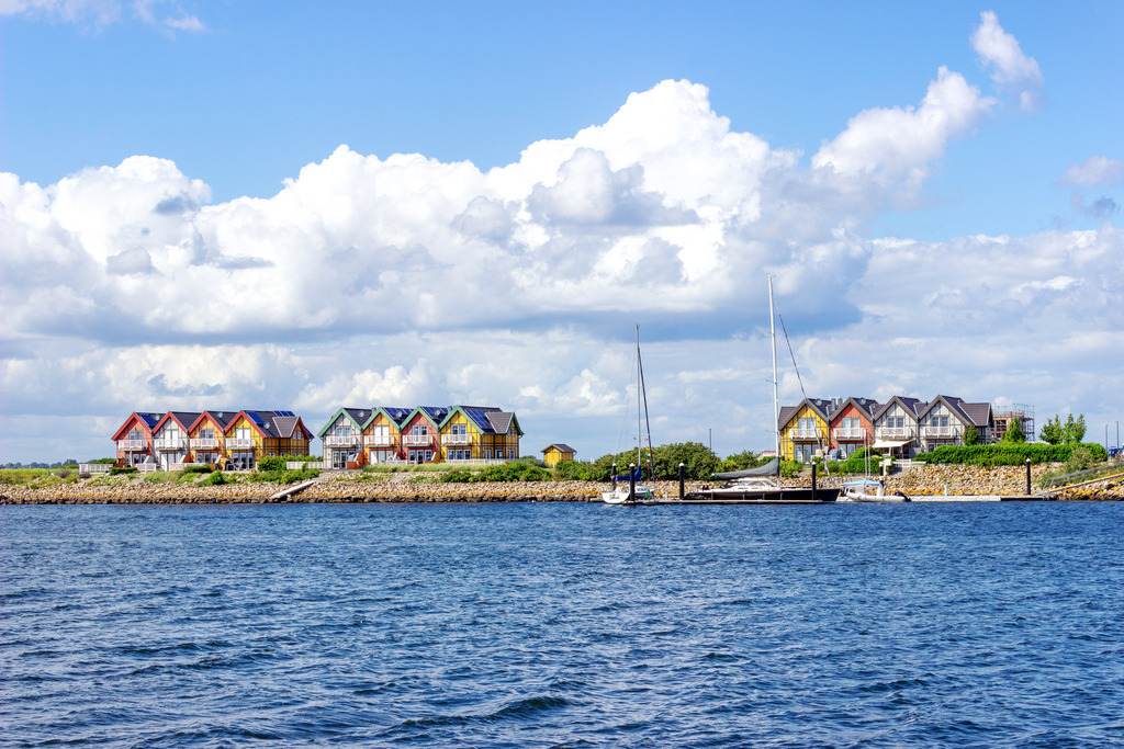 Wandbild: Malerische Ferienhäuser am Meer in Olpenitz | Dieses Wandbild im Querformat zeigt bunte Ferienwohnungen direkt am Meer in Olpenitz.Vor den Ferienwohnungen liegen zwei Segelboote. Im Vordergrund ist das Meer zu sehen. Am Himmel befinden sich sommerliche Wolken.  - Realisiert mit Pictrs.com