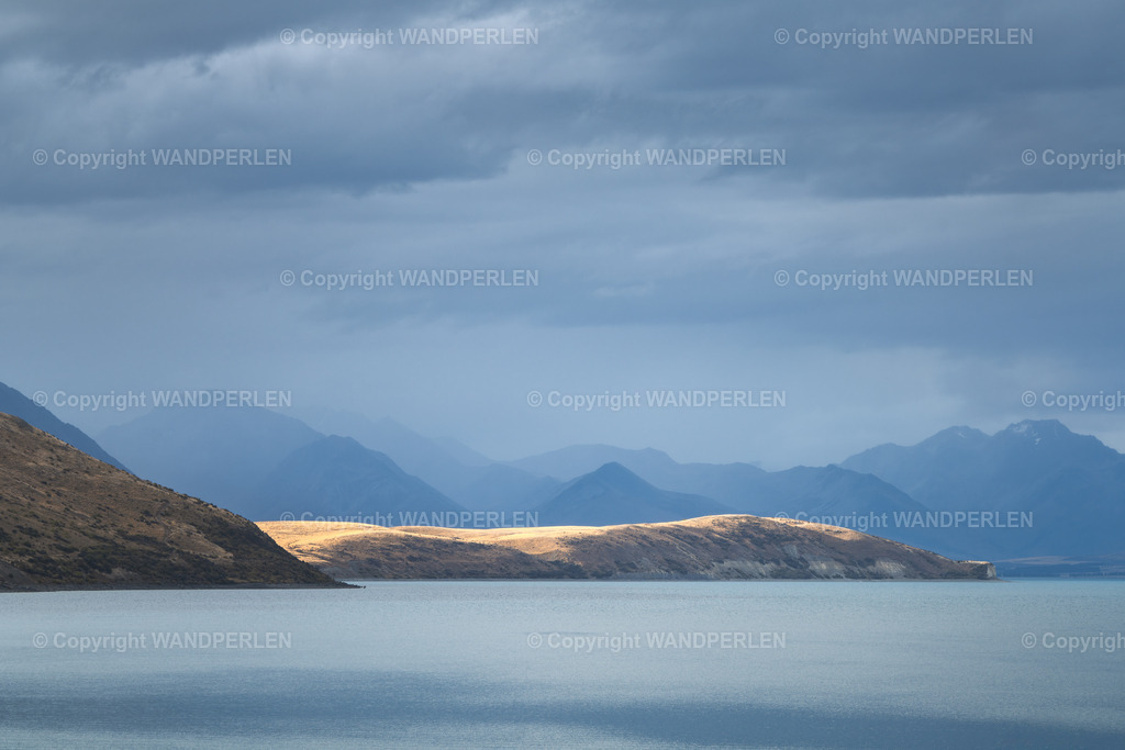 Lake Tekapo | Eine dramatische und atmosphärische Szene am See von Tekapo. Dunkle, bedrohliche Wolken hängen über der ikonischen Bergkette. Die Oberfläche des Sees bleibt ruhig und glatt. Die Hügel im Vordergrund sind in ein warmes, goldenes Licht getaucht, was einen auffälligen Kontrast zu den kühlen, gedämpften Tönen der entfernten Gipfel und des bewölkten Himmels schafft. Dieses Bild unterstreicht die mächtige und ständig wechselnde Natur der Landschaft der Südinsel Neuseelands. - Realisiert mit Pictrs.com