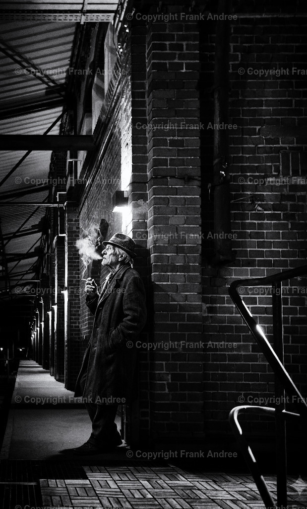 Am Ende des Tages | Ein älterer Mann in einem langen Mantel und Hut lehnt an einer Backsteinmauer, raucht eine Zigarette und ist in Gedanken versunken. Das Schwarz-Weiß-Foto fängt die Atmosphäre eines alten Film Noir perfekt ein – der dramatische Schein einer Wandlampe, der aufsteigende Rauch und der scheinbar endlose Bogen der Arkade im Hintergrund verleihen der Szene eine zeitlose, melancholische Stimmung. - Realisiert mit Pictrs.com