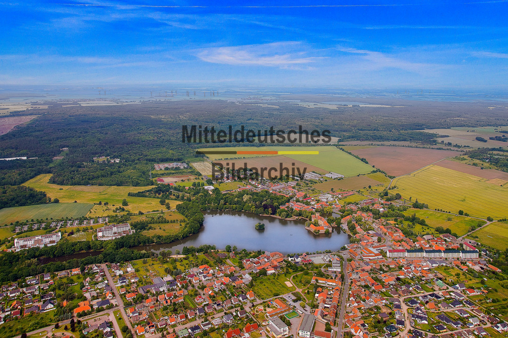 Flechtingen-Wasserschloß-Luftbild-Börde_gelb_3728 | Die Luftaufnahme zeigt Flechtingen mit dem See als prägendem Bestandteil des Ortsbildes. Wohnbebauung, Grünflächen und Wasser bilden aus der Vogelperspektive eine charakteristische Einheit. In der weiteren Umgebung sind außerdem Gewerbeflächen und der Steinbruch zu erkennen, die eine andere Seite der Region dokumentieren. - Realisiert mit Pictrs.com