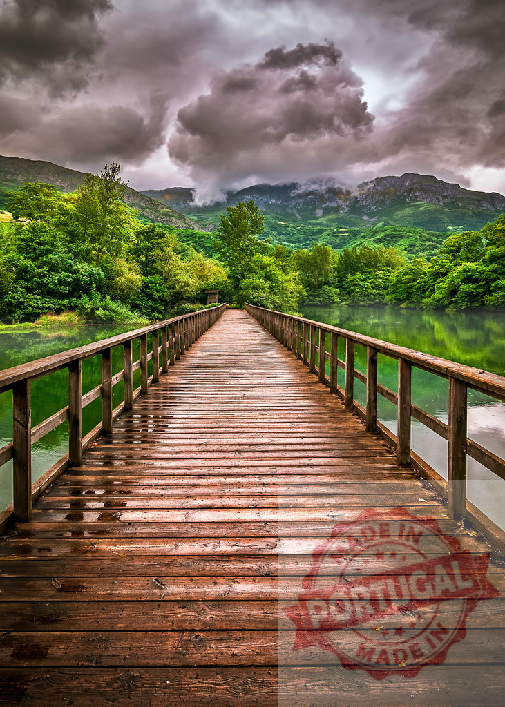 Wooden footbridge crossing a lake under a stormy sky | Wet wooden walkway over a lake leading to green hills and mountains under dark clouds - Realized with Pictrs.com