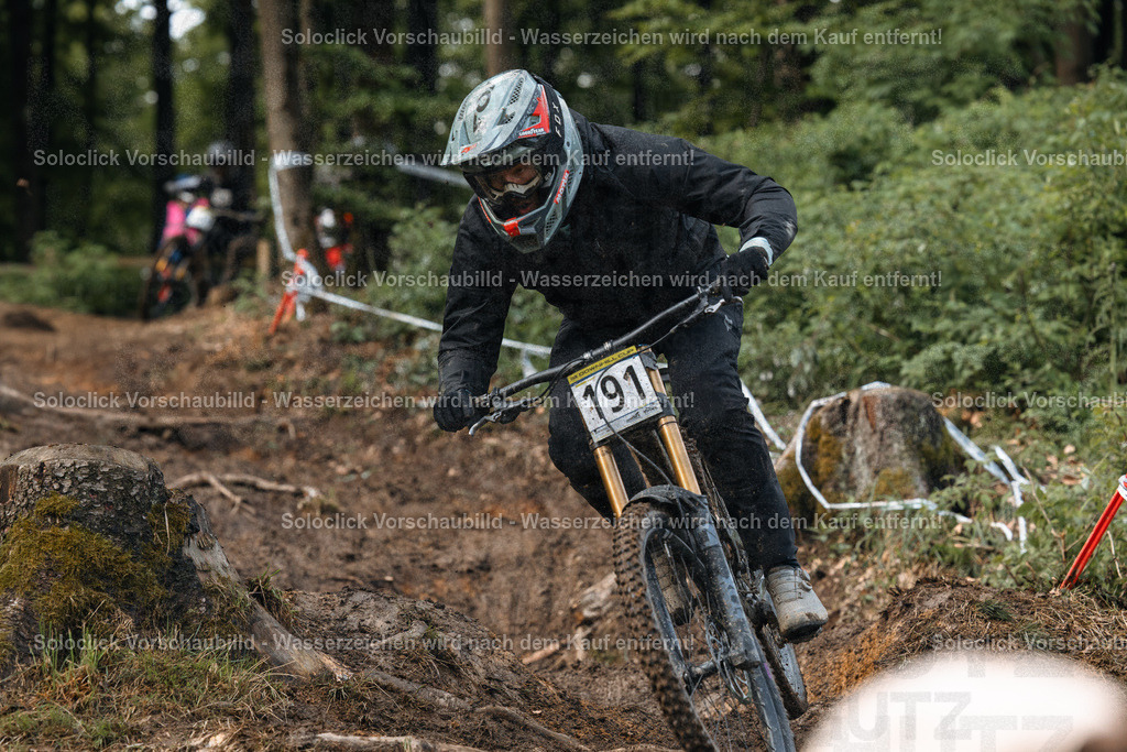 Mountainbike-Action im Wald | Ein Mountainbiker in voller Ausrüstung rast einen staubigen, kurvigen Trail hinunter. Er trägt einen schwarzen Overall, einen schwarzen Helm mit einer roten und weißen Grafik und eine Nummer 191 auf seinem Helm und Trikot. Der Trail schlängelt sich durch einen dichten Wald, der mit Bäumen und Wurzeln übersät ist. Im Hintergrund sind weitere Mountainbiker zu sehen, die ebenfalls den Trail befahren. Die Szene vermittelt ein Gefühl von Geschwindigkeit, Abenteuer und Herausforderung. - Realisiert mit Pictrs.com