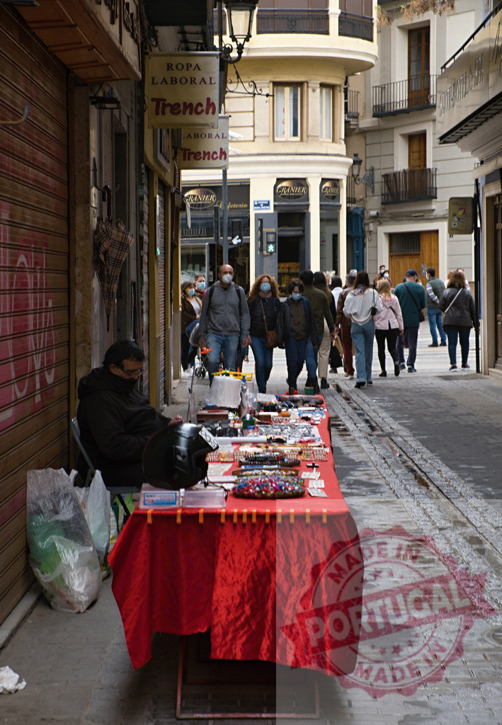 Man selling items on a busy city street in Valencia | Man selling various items on a red table with people walking past on a narrow city street - Realized with Pictrs.com