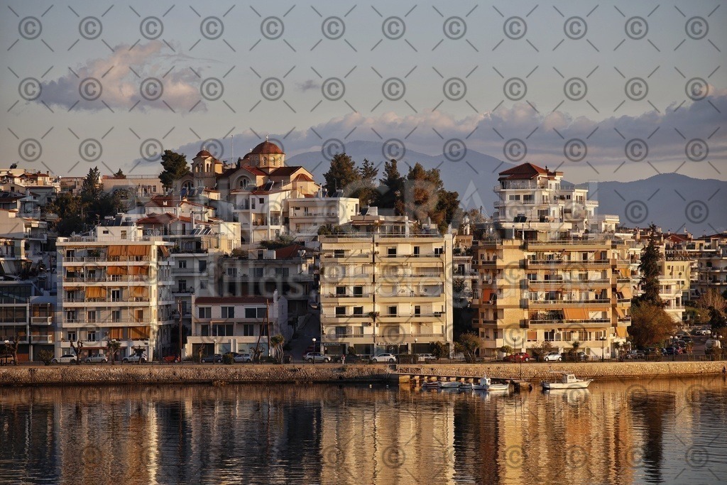 Die Skyline von Chalkida | Die Skyline der Festlandseite von Chalkida (Χαλκίδα). Chalkida ist die Hauptstadt der der griechischen Insel Euböa (Εύβοια). Chalkida erstreckt sich über beide Seiten der Meeresstraße Euripos (Εύριπος), welche zwischen dem griechischen Festland und der Insel Euböa liegt. 
Über den Dächern der Promenade ragt die Kuppe der griechisch-orthodoxen Kirche "Ekklisia Evaggelismos Theotokou" (Εκκλησία Ευαγγελισμός Θεοτόκου). Im Vordergrund spiegelt sich die Skyline im Morgenlicht in der Meeresenge von Euripos. Die Meerenge ist an der engsten Stelle nur 40 Meter breit und gilt als schmalste Meerenge der Welt. Eine Besonderheit der Meerenge sind die wechselnden Gezeitenströmungen. Die regulären Gezeitenströmungen wechseln alle 6 Stunden die Richtung. Zwei mal pro Mondmonat treten die irregulären Strömungen auf, die bis zu vierzehn mal am Tag die Richtung wechseln.