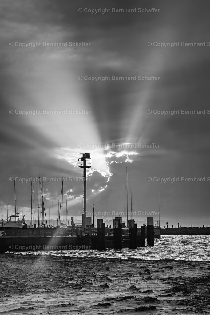 Landungssteg im Sonnenlicht | Ein Sandstrand an der Kieler Förde und ein Steg mit Sonnenstrahlen, die nach einem Gewitter durch die Wolken brechen, schwarz-weiss. - Realisiert mit Pictrs.com