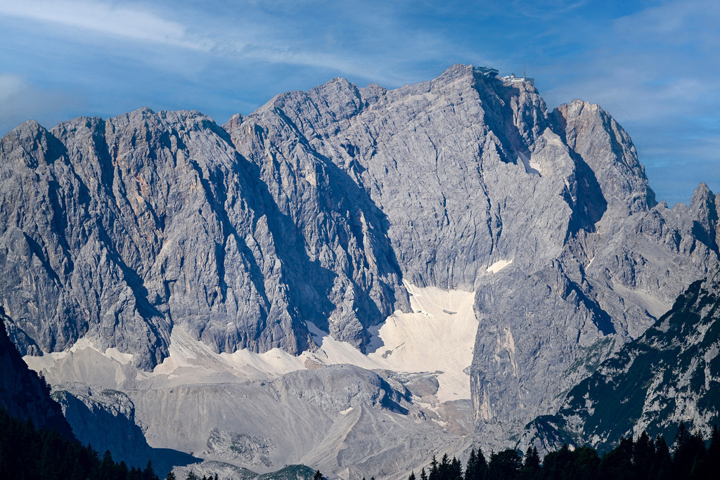 Thron der Alpen: Die urgewaltige Pracht der Zugspitze | Ein Anblick von atemberaubender, roher Monumentalität, der tiefe Ehrfurcht vor den Kräften der Natur weckt: Stolz und unbezwingbar ragt das gewaltige Massiv der Zugspitze empor. Die schroffen, tief zerfurchten Felswände aus grauem Kalkstein offenbaren jede einzelne Falte jahrtausendealter Erdgeschichte, während in den tiefen Karen letzte, strahlend weiße Schneefelder und Reste des Gletschers trotzen. Weit oben auf dem höchsten Gipfel Deutschlands thront die markante Bergstation als winziges Zeugnis menschlicher Pionierarbeit. Diese Aufnahme fängt die erhabene Einsamkeit, die gewaltige Dimension und die zeitlose Faszination des Hochgebirges perfekt ein. - Realisiert mit Pictrs.com