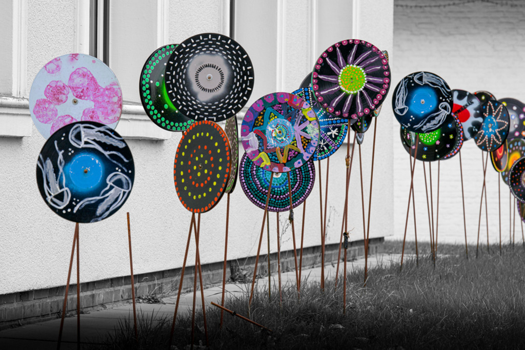 Schallplatten-Haus | Bunt bemalte Schallplatten sind an rostigen Stahlstäben befestigt, stehen aufgereiht vor einem Haus. Die Farben wurden auf die Schallplatten und Stahlstäbe reduziert, das Umfeld ist schwarz-weiß. — Auflösung des Originals: 8256 x 5504 px. - Realisiert mit Pictrs.com