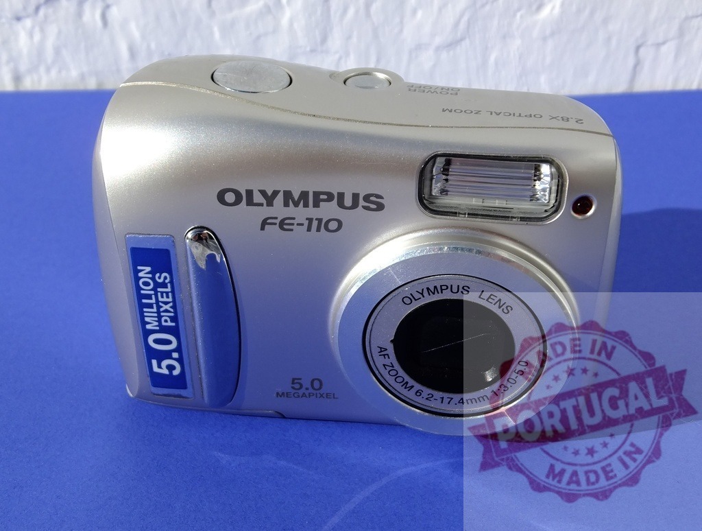 Olympus FE-110 vintage digital point and shoot camera | Olympus FE-110 silver digital camera on a blue background, representing retro technology - Realized with Pictrs.com