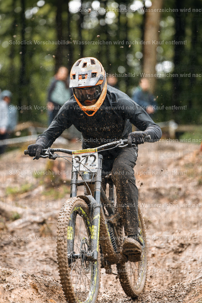 Adrenalin im Wald | Ein Mountainbiker in voller Ausrüstung rast auf einem staubigen Pfad durch einen dichten Wald. Er trägt einen schwarzen Helm mit orangefarbener Visier, eine schwarze Jacke und Handschuhe. Sein Mountainbike ist mit breiten Reifen und einer robusten Federgabel ausgestattet. Staub wirbelt hinter ihm auf, was die Geschwindigkeit und die Action des Moments unterstreicht. - Realisiert mit Pictrs.com