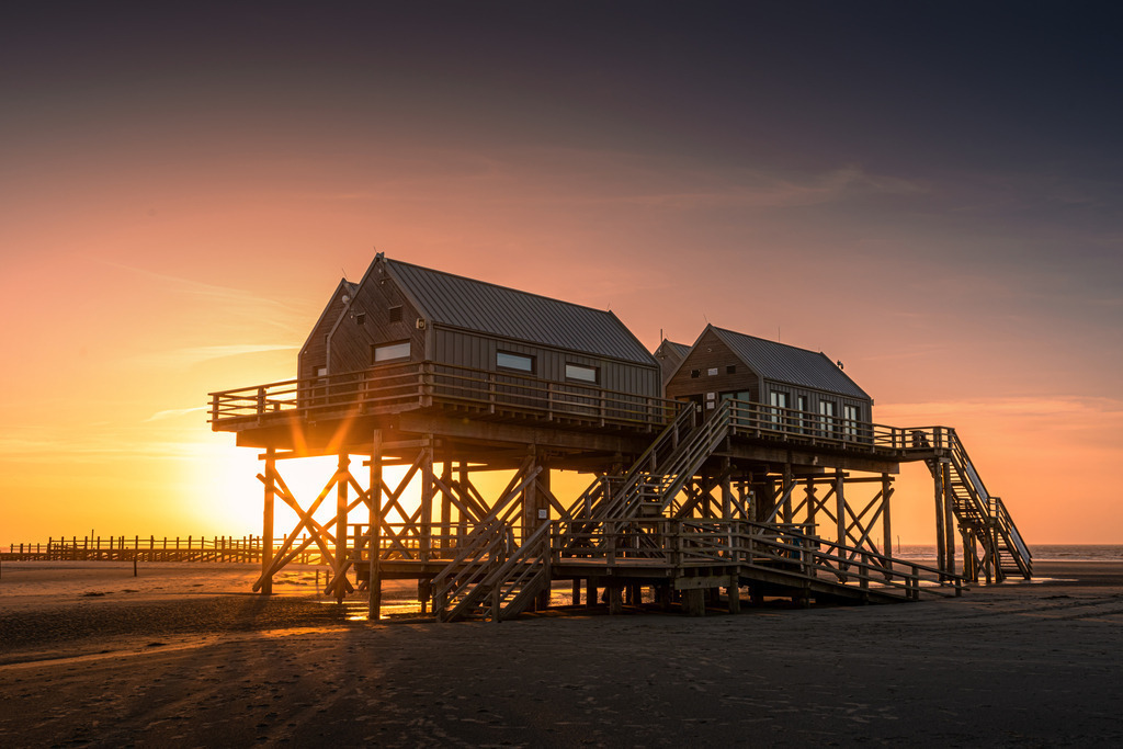 Sonnenuntergang in St. Peter Ording | Auf dem Bild sind die charakteristischen Pfahlbauten von St. Peter-Ording bei Sonnenuntergang zu sehen. Die Holzhäuser stehen auf hohen Stelzen, um sie vor Hochwasser zu schützen. Sie sind über Holztreppen und -plattformen zugänglich und bieten einen eindrucksvollen Blick auf das Meer und den Strand. Die warmen Sonnenstrahlen tauchen die Szene in ein stimmungsvolles, goldenes Licht.