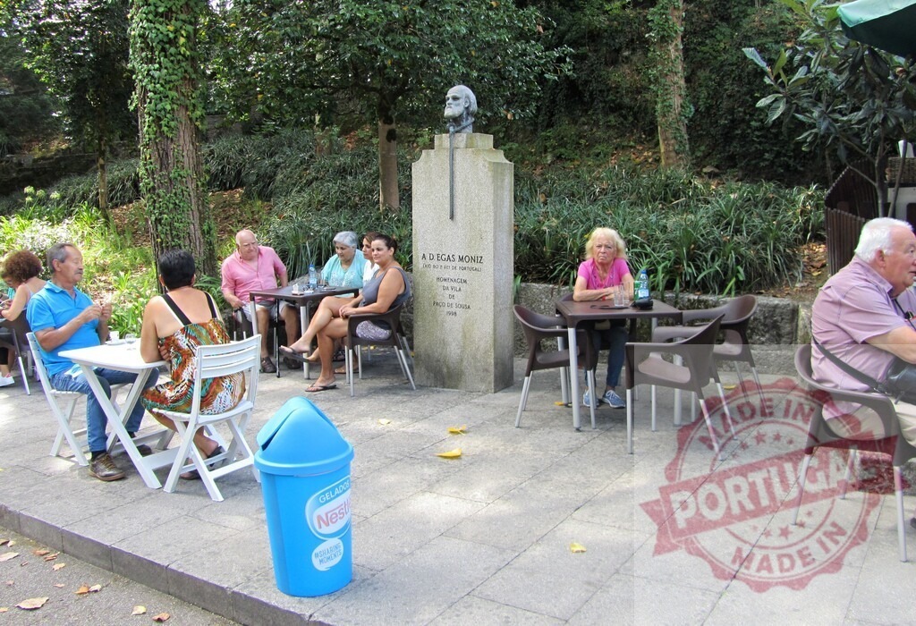 People enjoying outdoor cafe seating near Egas Moniz bust in Portugal | People relaxing at an outdoor cafe beside the Egas Moniz bust on a sunny day - Realisiert mit Pictrs.com