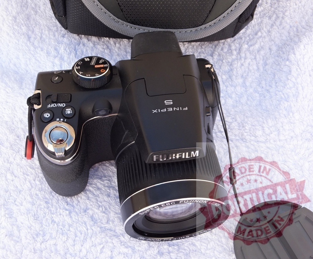 Fujifilm FinePix digital bridge camera on white towel | Fujifilm FinePix digital camera with lens cap on a white fabric surface with a camera bag - Realized with Pictrs.com