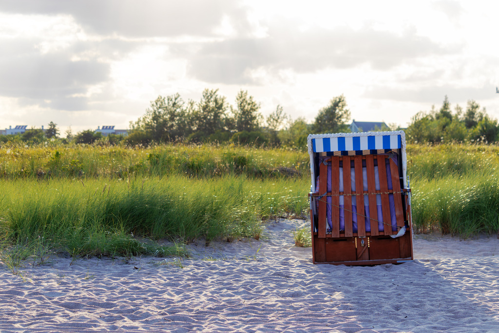 Wandbild: Strandkorb am Strand in Weidefeld | Dieses Wandbild im Querformat zeigt einen charmanten Strandkorb am Strand von Weidefeld. Die sommerlichen Strandgräser im Hintergrund und der teils bewölkte Himmel verleihen der Szene eine ruhige und einladende Atmosphäre. Holen Sie sich dieses maritime Kunstwerk in Ihr Zuhause und genießen Sie die entspannende Wirkung. - Realisiert mit Pictrs.com