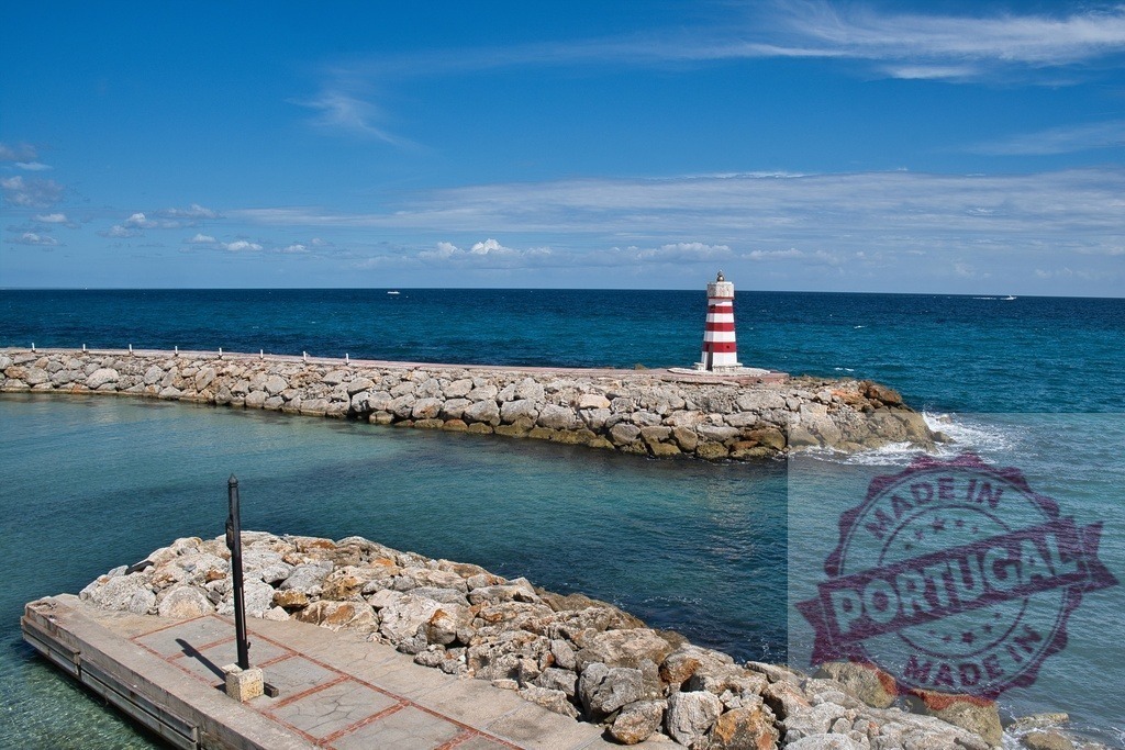 Red and white striped lighthouse guiding clear ocean waters | Red and white striped lighthouse sitting on a rocky pier by the turquoise ocean under blue sky - Realized with Pictrs.com