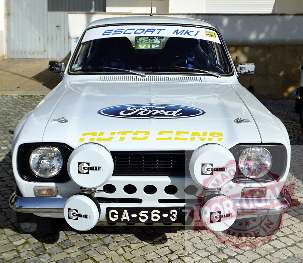 White Ford Escort MKI Rally Car Parked on Cobblestone Street | Classic white Ford Escort MKI rally car parked, showing front view with Cibie spotlights and Auto Sena branding - Realized with Pictrs.com