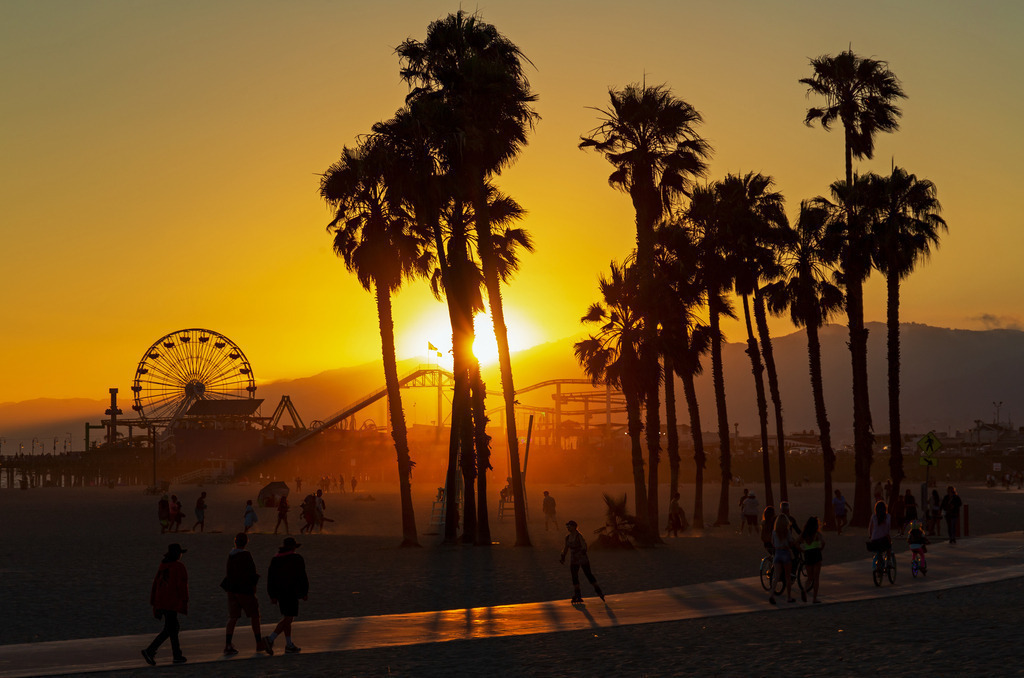 Wandbild - Sonnenuntergang am Venice Beach | Dieses stimmungsvolle Bild fängt den Sonnenuntergang am berühmten Venice Beach in Los Angeles ein. Die Silhouetten der hohen Palmen ragen vor dem leuchtend orangefarbenen Himmel empor, während die untergehende Sonne hinter den Santa Monica Mountains verschwindet. Im Hintergrund sind das Riesenrad und die Achterbahnen des Santa Monica Piers zu sehen, die der Szene ein nostalgisches Flair verleihen. Menschen spazieren, skaten und radeln entlang der Promenade, was das lebendige und entspannte Ambiente von Venice Beach unterstreicht.