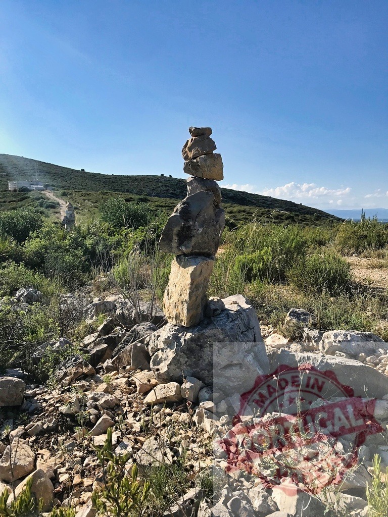 Stone cairn balancing rocks on a hiking trail | Cairn of balanced stones marking a path on a rocky, scrubland hillside under a clear blue sky - Realized with Pictrs.com