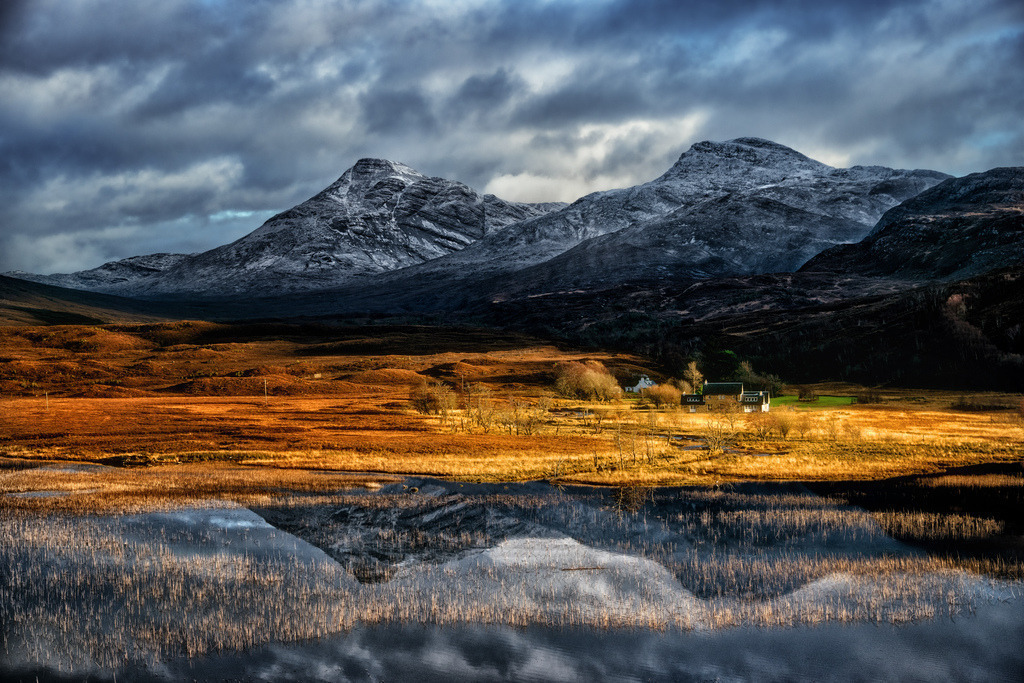 Winterlandschaft am Loch Damh | Eine weite schottische Winterlandschaft erstreckt sich mit einem See im Vordergrund, der die schneebedeckten Berge und den dramatischen Himmel widerspiegelt. Goldbraune Moorlandschaft wird von tiefstehender Sonne beleuchtet, während dunkle Wolken über den Gipfeln hängen. Einige Gebäude am Ufer fügen einen menschlichen Akzent in die unberührte Natur ein. - Realisiert mit Pictrs.com