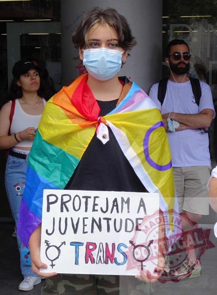 Person holding Protejam a Juventude Trans sign at Pride protest | Young person wearing a face mask and a Pride flag holding a sign for trans youth protection - Realisiert mit Pictrs.com