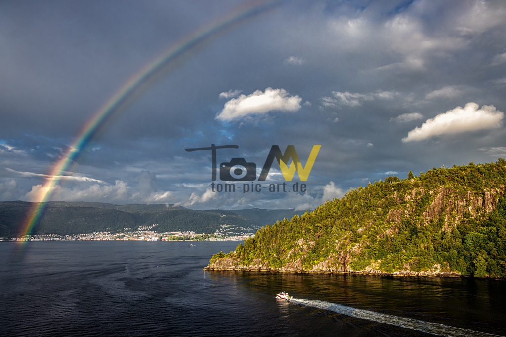 Einfahrt in den Hafen von Bergen--Regenbogen------Norwegen | Die Aufnahme fängt einen natürlichen Moment ein, in dem ein heller Regenbogen unter einem bewölkten Himmel auftaucht und das Wasser berührt. Im Vordergrund ist ein bewaldeter Hügel zu sehen, während im Hintergrund eine Stadt entlang der Küste liegt. Ein kleines Boot hinterlässt eine Spur auf dem dunklen Wasser.Bergen ist bekannt als das Herz der norwegischen Fjorde und bietet eine Kombination aus Natur und Kultur.  - Realisiert mit Pictrs.com