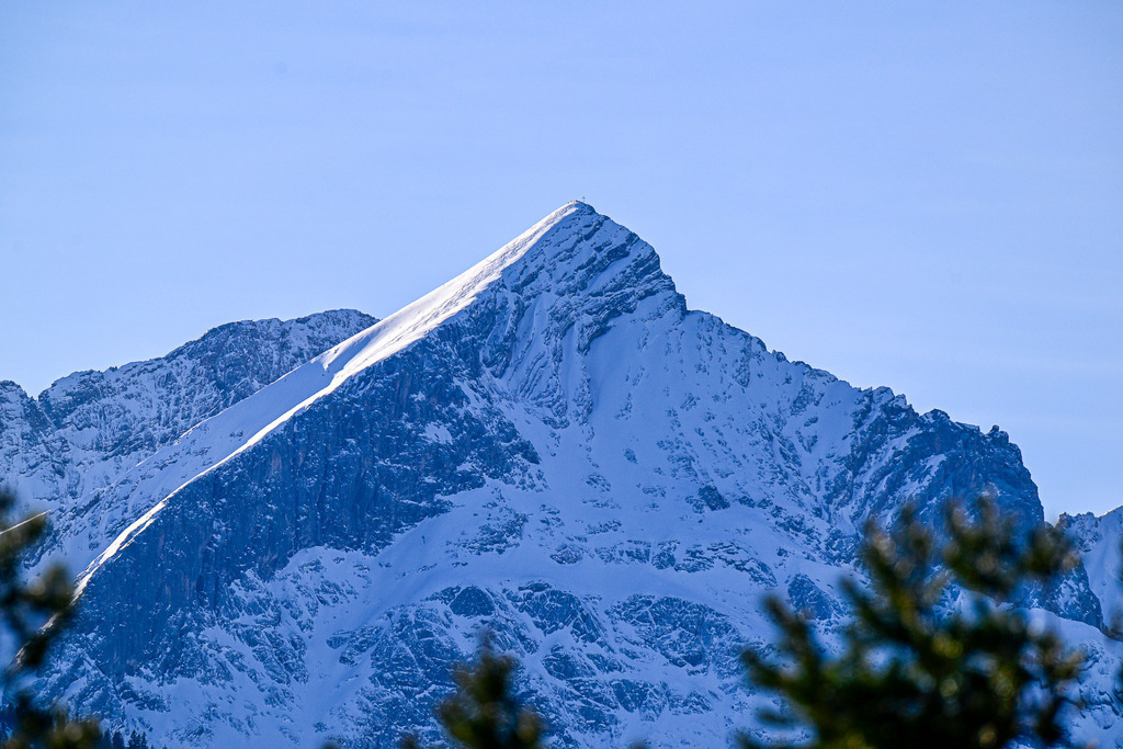 Winterliche Krone der Alpen: Die verschneite Alpspitze | Ein eiskaltes, erhabenes Kunstwerk der Natur: Stolz und unnahbar ragt die markante Pyramide der Alpspitze in den makellos blauen Winterhimmel. Auf der messerscharfen Krete funkelt der frisch gefallene Schnee im strahlenden Sonnenlicht, während die steilen Nordwände in mystisches, kühles Schattenblau getaucht sind. Am äußersten Gipfelpunkt wacht das winzige Kreuz über der unendlichen Stille. Gerahmt von dunklen Tannenzweigen im Vordergrund, weckt dieser majestätische Anblick eine tiefe Sehnsucht nach der reinen, unberührten Freiheit der winterlichen Bergwelt. - Realisiert mit Pictrs.com